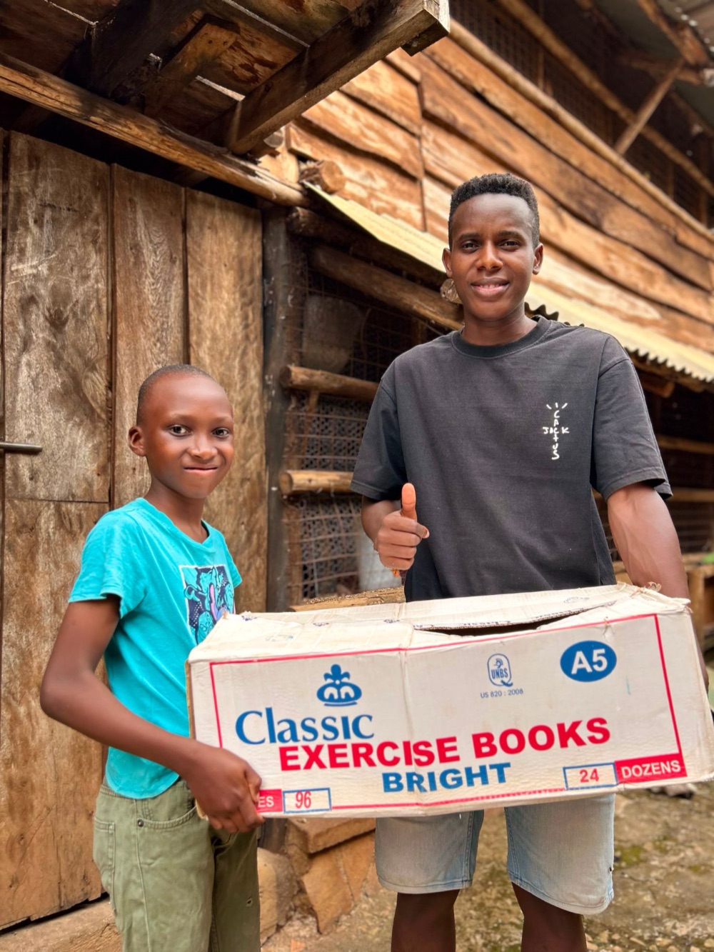 A boy and a young man giving a thumbs up behind a box of exercise books