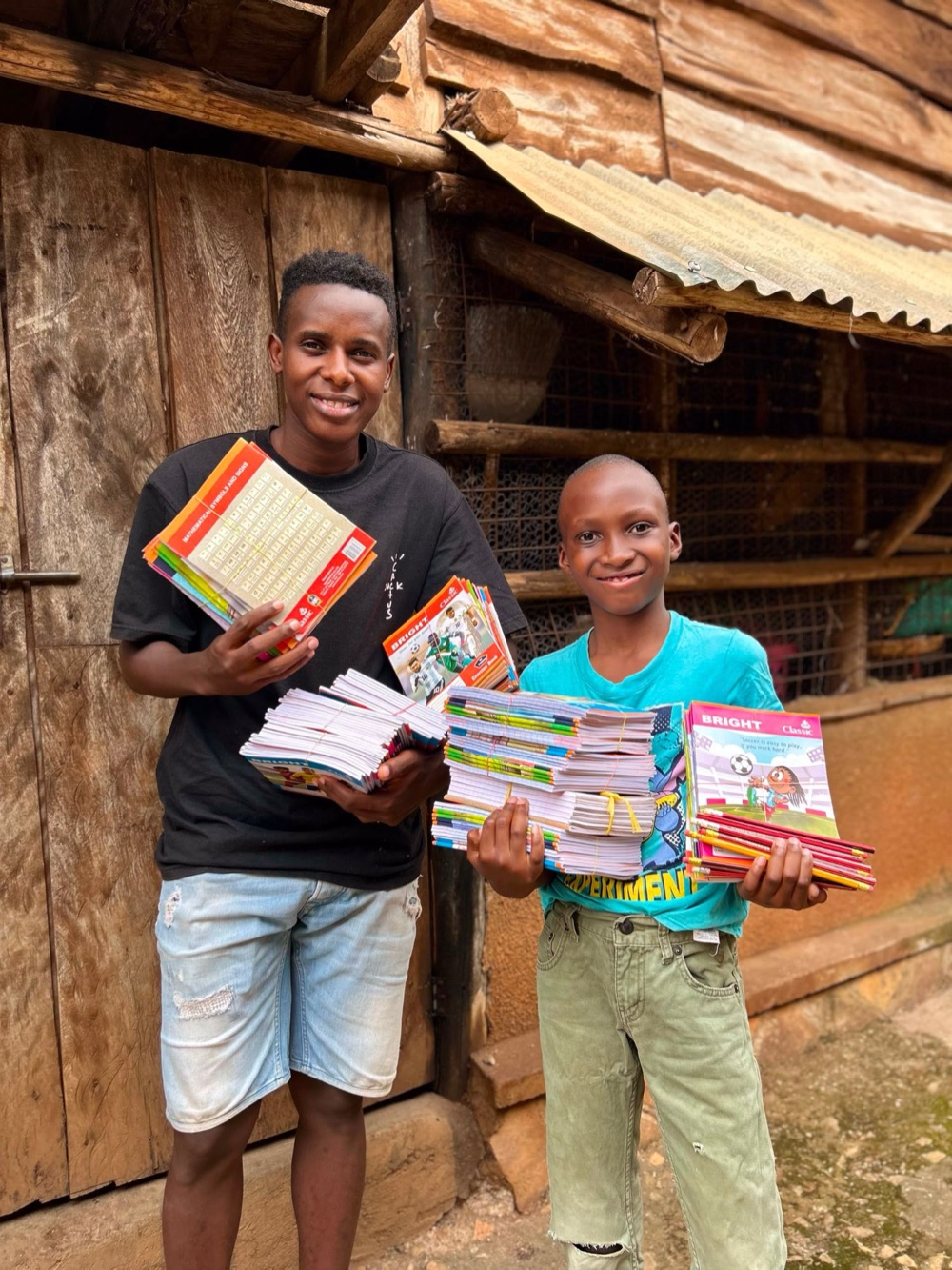 A young man and a boy smiling outside a wooden building, arms full of exercise books and pencils