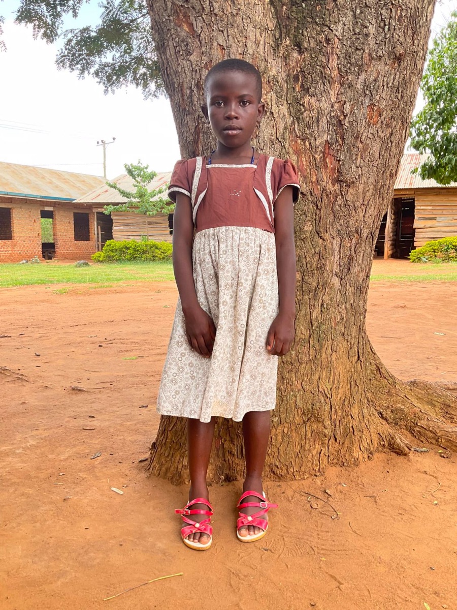 A girl in a brown and cream dress and red sandals standing in front of a large tree