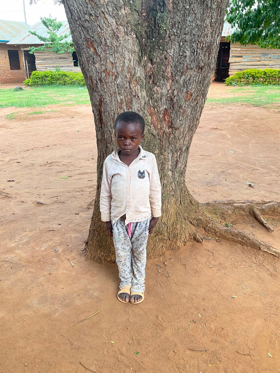 A small child in a pale shirt and printed trousers standing in front of a large tree