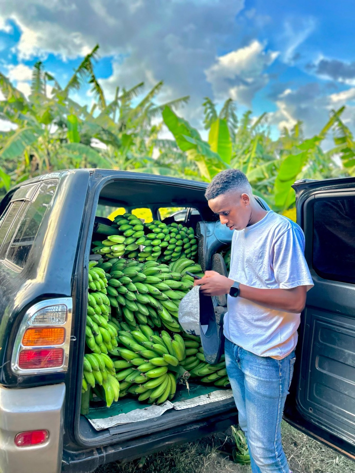 A young man beside a car whose boot is packed with green matooke bunches, banana plants behind
