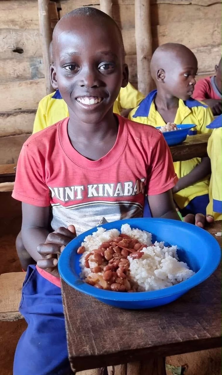 A smiling boy in a red T-shirt with a bowl of rice and beans at his school desk