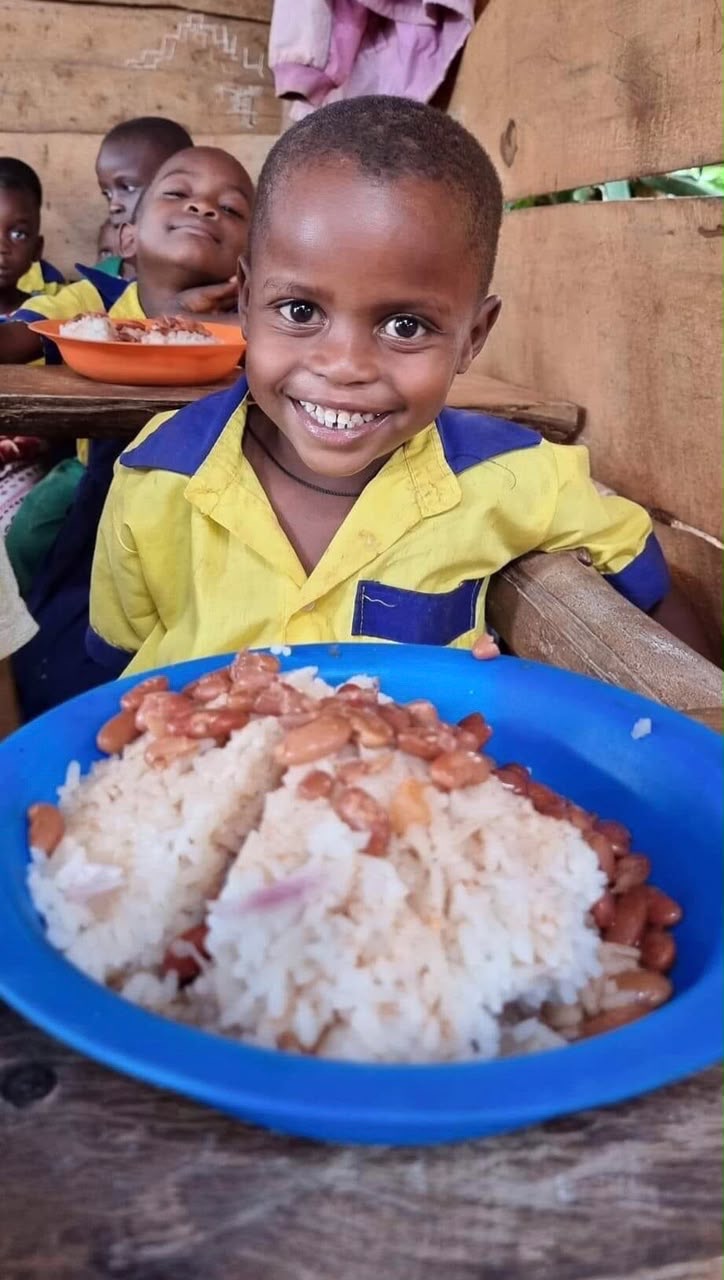 A small child in a yellow school shirt smiling over a big bowl of rice and beans