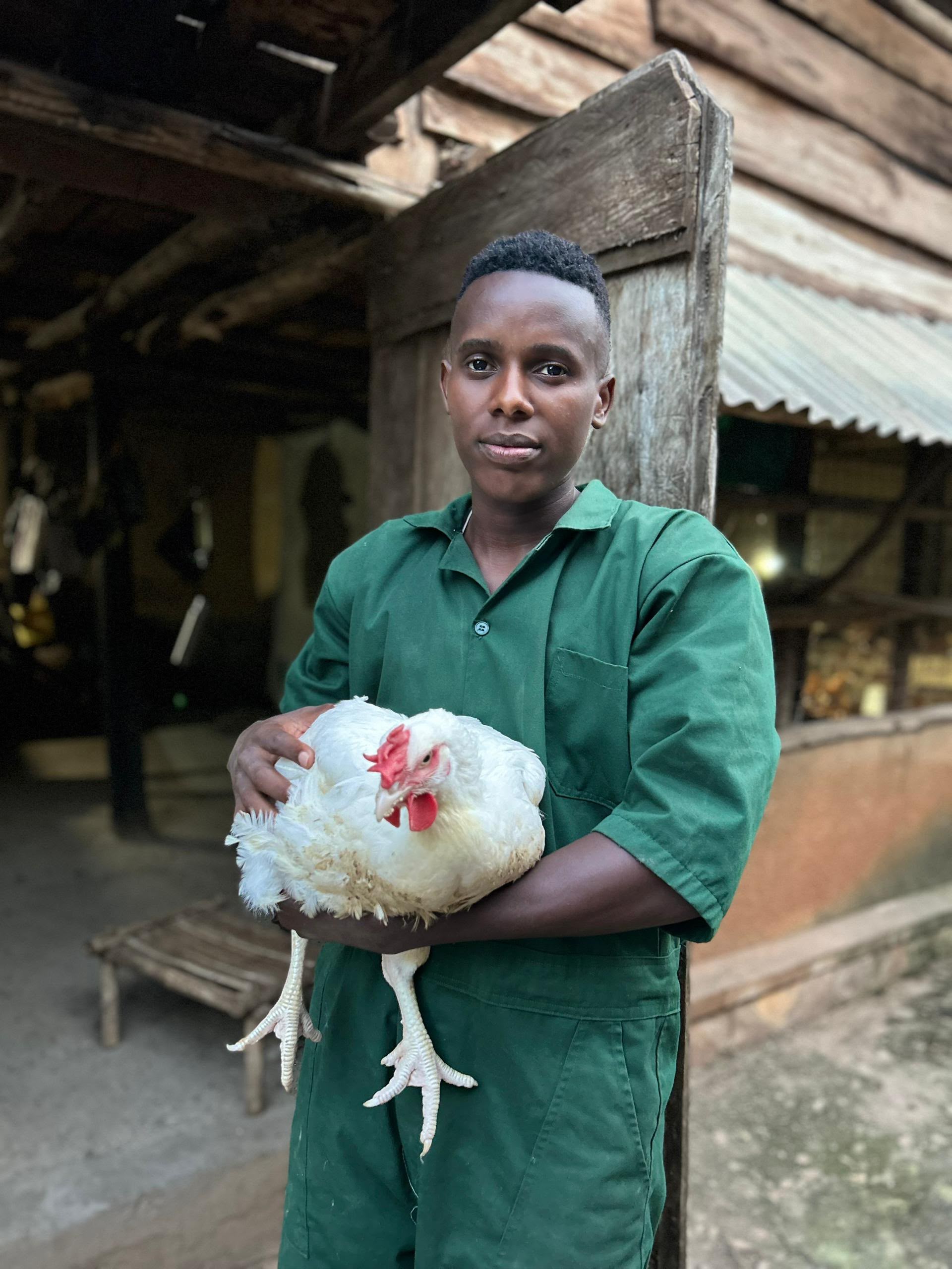 Thomas Turahe in green overalls, holding a white hen outside a wooden farm building