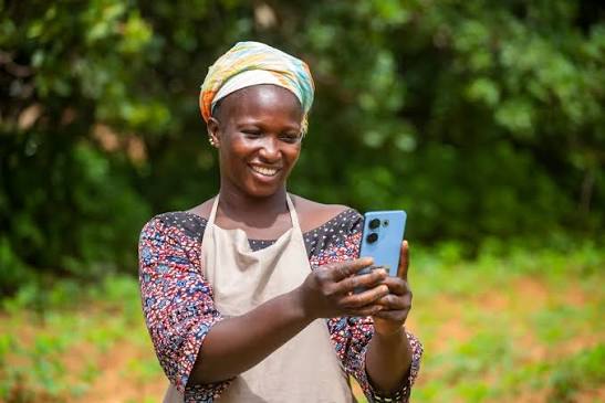 A smiling woman in a headscarf and apron holding up a phone on a video call outdoors