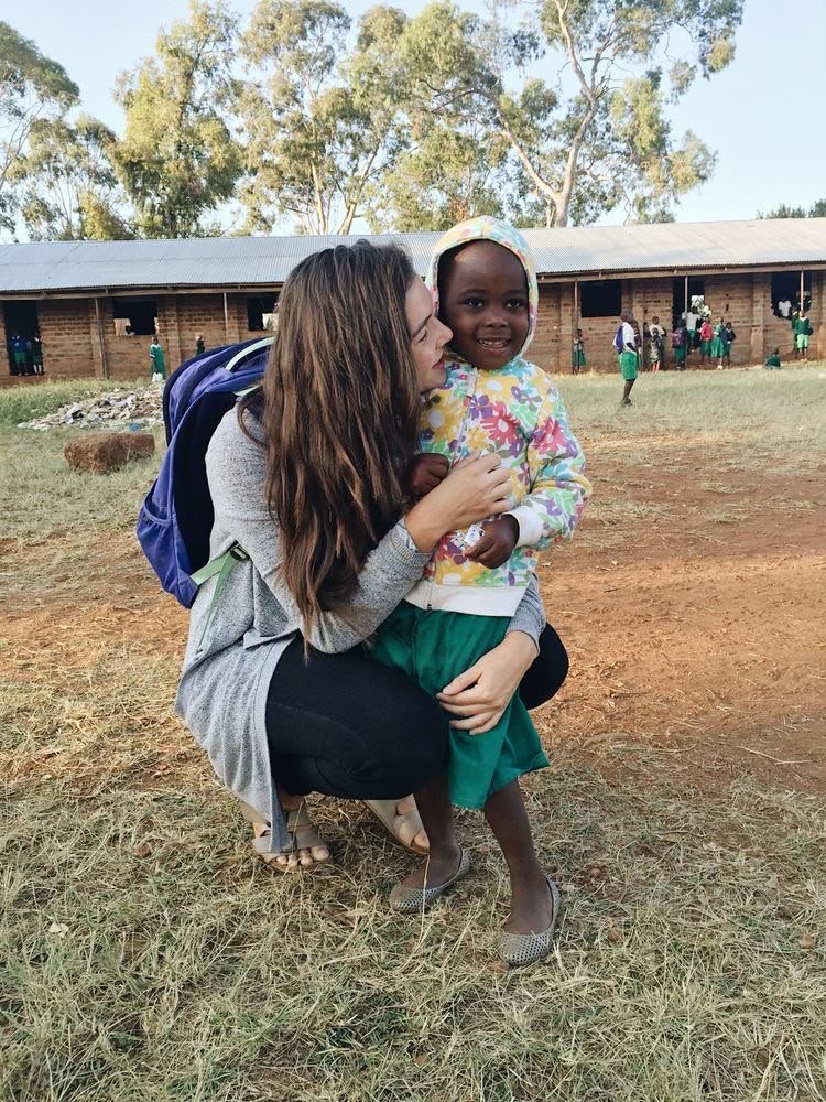 A volunteer crouching to hug a smiling little girl in a flowered hoodie outside a school