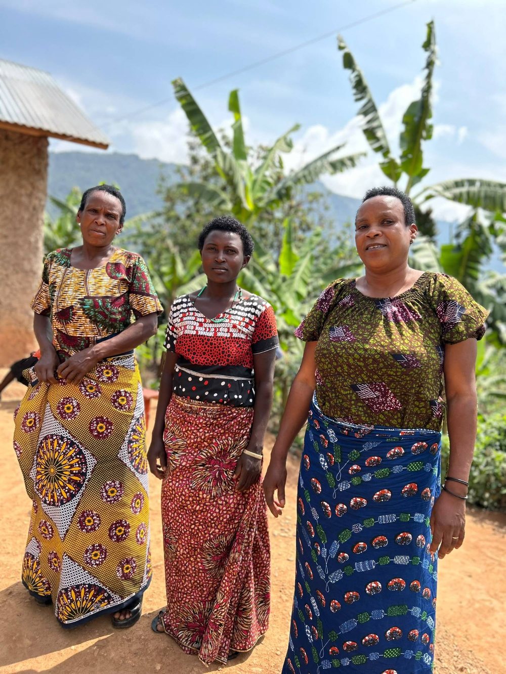 Three women from the collective in bright printed wraps standing on a red earth path, banana plants and hills behind