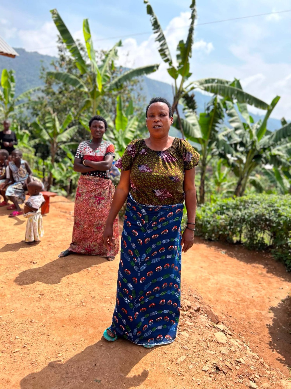 A woman in a blue patterned wrap speaking to the group, with banana plants and hills behind her