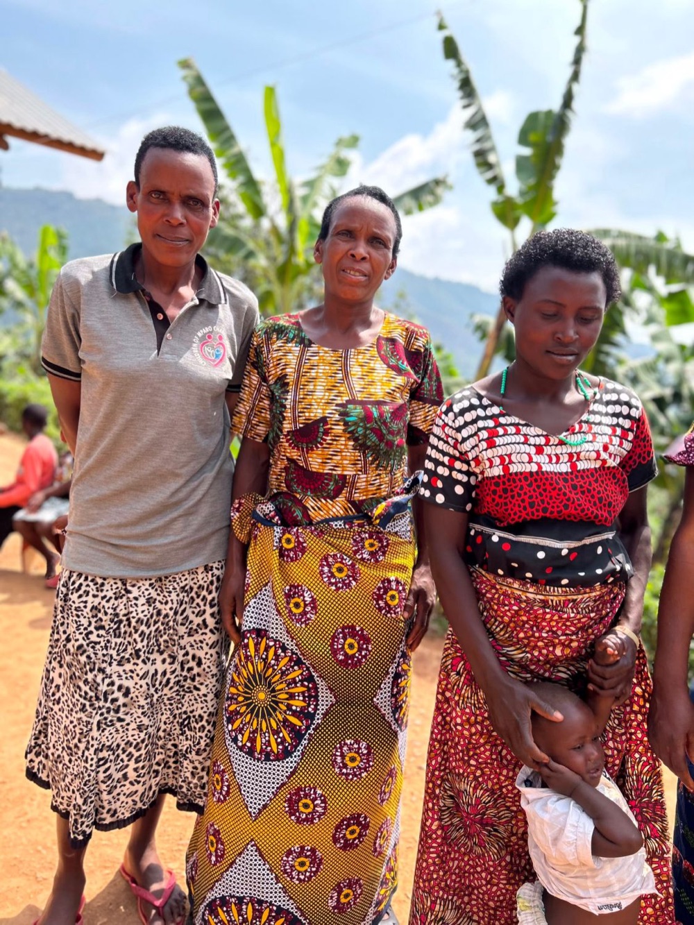 Three women in bright printed dresses standing together among banana plants